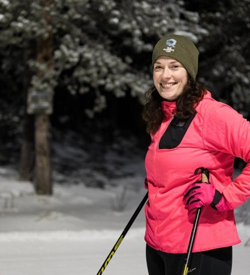Woman wearing bright pink jacket and black training pants standing on cross country skis on dark forested ski trail