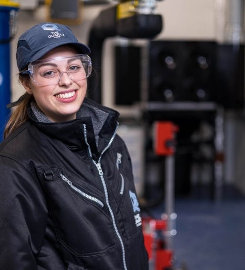 Young woman in hard hat, glasses and black coat standing in a factory smiling at camera